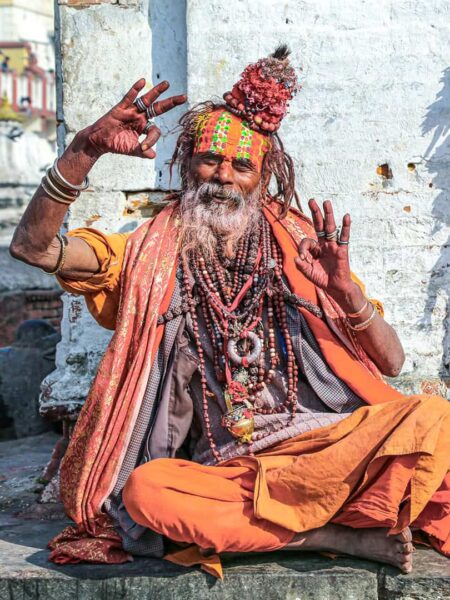 Man sieht einen Sadhu in Kathmando. Er hat einen langen, grauen Bart und trägt orange und Gelbe Kleidung sowie Schmuck aus Holzperlen. Er sitzt im Schneidersitz, hat beide Hände halb gehoben und lächelt schief in die Kamera.