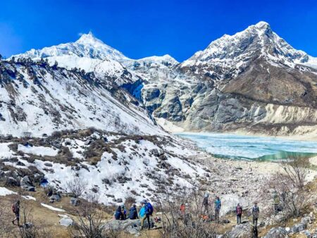 Mehrere Wanderreisende in einer kleinen Gruppe gehen auf den Birendra-See in Nepal zu. Der See erstrahlt in sehr hellem Blau und rundherum liegt ein wenig Schnee auf dem sonst recht kahlen Fels. Im Hintergrund sieht man zu Teilen schneebedeckte Berge.
