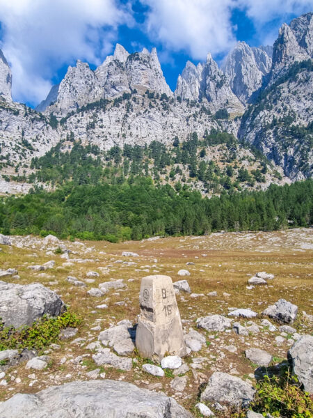 Ein Grenzstein, der die Grenze von Albanien und Montenegro markiert. Er steht allein auf einem Feld und kann problemlos im Zuge einer Wanderung passiert werden.