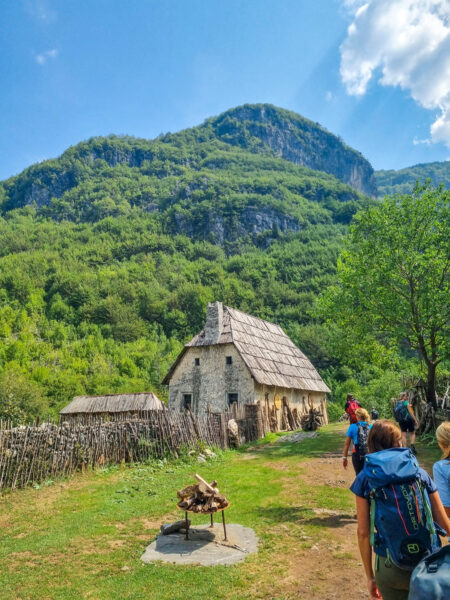 Ein albanisches Haus, das am Weg von oder zum Wasserfall von Teth am Wegesrund zu finden ist. Mehrere Personen auf Wanderreise gehen gerade daran vorbei.