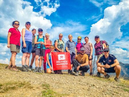 Auf dem Bild sieht man eine Wandergruppe auf einer Alm in den albanischen Alpen. Die Tourist:innen unterschiedlichen Alters stehen zum Gruppenbild beisammen und halten zwischen sich eine Flagge mit dem Logo des Reisenveranstalters Weltweitwandern.