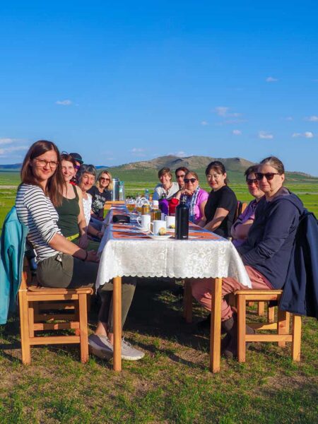 Eine Reisegruppe auf einer Wanderreise durch die Mongolei sitzt gemeinsam an einem gedeckten Tisch auf einer grünen Wiese in den Bergen der Mongolei.