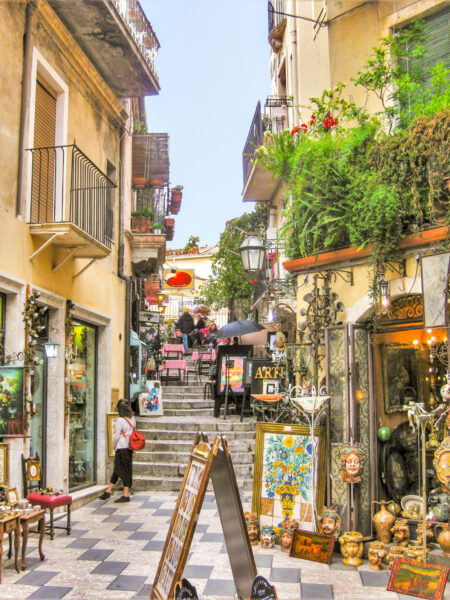 Das Bild zeigt eine Gasse der Ortschaft Taormina in Sizilien. Im Vordergrund sieht man eine Kaffee sowie einen Laden mit Antiquitäten. Im Hintergrund sieht man eine Person auf Wanderreise, die die Stufen einer Treppe in der Altstadt erklimmt.