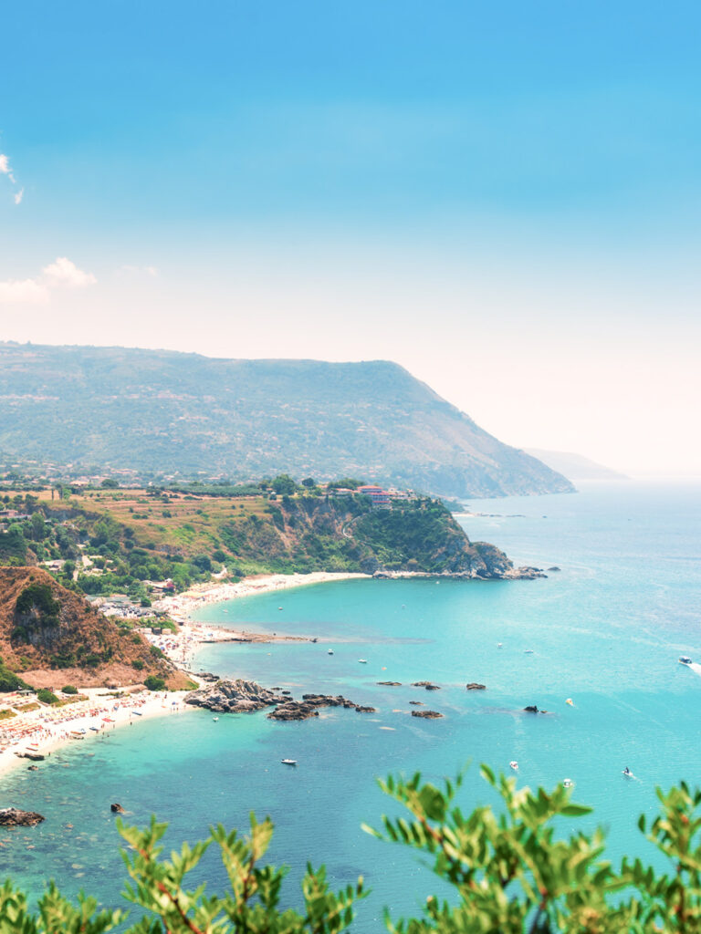Die Küste von Italien in der Nähe der Stadt Tropea ist im Bild zu sehen. Man sieht das strahlend-blaue tyrrhenische Meer über die verzeinzelten Büsche entang eines Wanderwegs hinweg.
