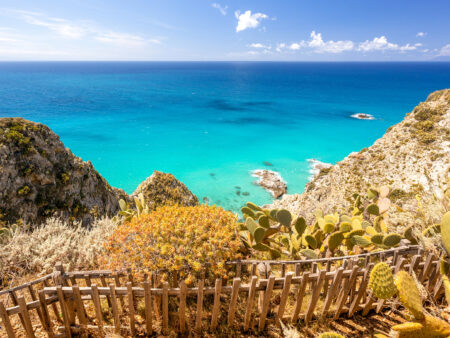 Ein Bild von der italienischen Küste am Capo Vaticano in Kalabrien. Zwischen dem Wanderpfad im Vordergrund und den steilen Klippen steht ein kleiner Holzzaun.