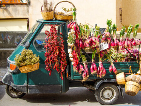 Ein kleiner, dreirädriger Lieferwagen steht am Straßenrand in Kalabrien. Überall von dem kleiner Wagen hängt getrocknetes oder auch frisches Gemüse sowie Töpfe mit diversen Kräutern.