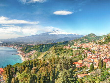 Der Ätna ragt in der Ferne in den Himmel auf. Im Vordergrund sieht man die Küste Siziliens, sowie ein Bergdorf, das halb im Wald auf der italienischen Insel versteckt liegt.