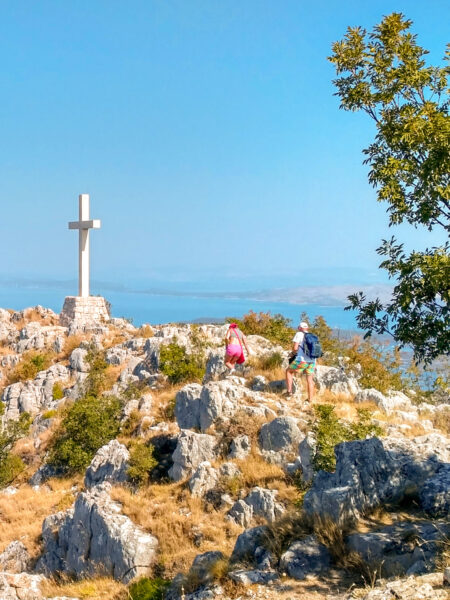 Ein kleines Gipfelkreuz auf einer kroatischen Insel. In der Ferne lässt sich der Ozean erkennen, mehrere Personen gehen auf ihrer Wanderreise auf das Gipfkreuz zu.