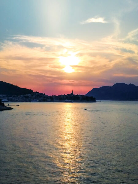 Die Sonne geht hinter der kroatischen Ortschaft Korčula unter. Sie reflektiert im Wasser der Bucht der kroatischen Insel.
