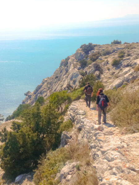 Zwei Personen auf Wanderreise gehen einen Weg entlang der schroffen Küste der Insel Brac in Kroatien entlang. Hinter ihnen sieht man die Adria leuchten.