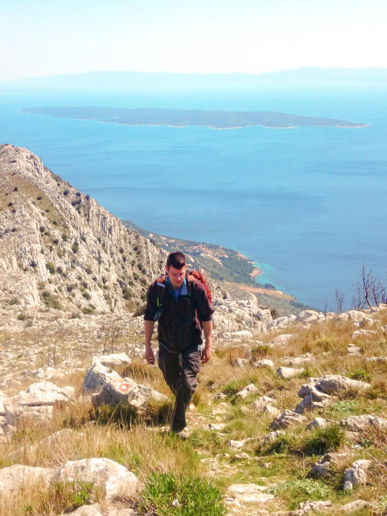 Ein Mann mit Wanderrucksack wandert einen Hang auf der Insel Hvar in Kroatien hinauf. Hinter ihm kann man von dem Berg, den er gerade hoch-trekkt, die Adria sehen.