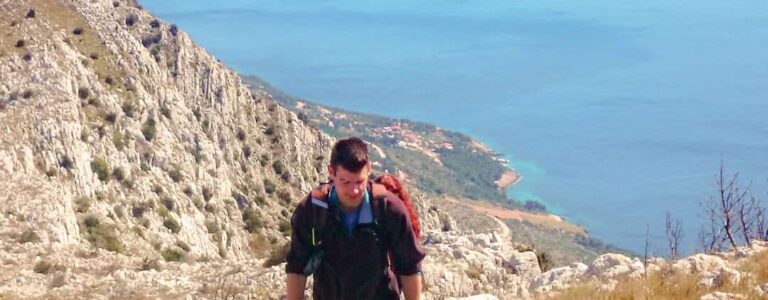 Ein Mann mit Wanderrucksack wandert einen Hang auf der Insel Hvar in Kroatien hinauf. Hinter ihm kann man von dem Berg, den er gerade hoch-trekkt, die Adria sehen.
