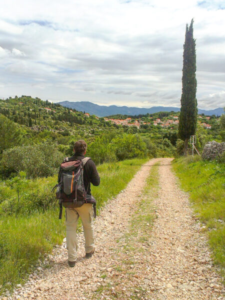 Eine Person mit Rucksack wandert einen Schotterweg auf der Insel Korčula in Kroatien entlang. In der Ferne sieht man eine Zypresse sowie eine kleine Ortschaft.
