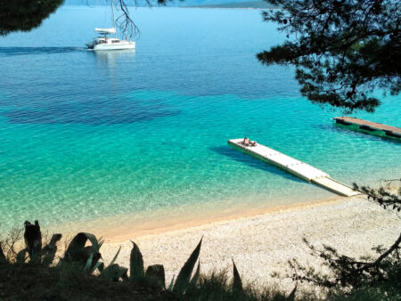 Ein kleines Boot liegt vor dem Sandstrand der Insel Brač, ein Steg führt vom Strand hinaus, wo sich zwei Personen auf Wanderreise gerade sonnen.
