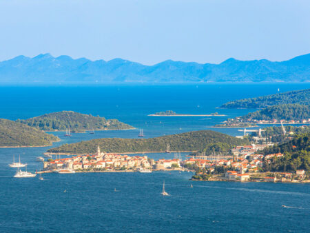 Die Stadt Korčula auf der gleichnamigen kroatischen Insel ist aus der Distanz fotografiert. Mehrere kleine Boote und Schiffe treiben im Hafen.