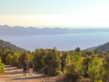Zwei Personen gehen einen breiten Wanderweg auf einer kroatischen Insel entlang. Im Hintergrund sieht man die Adria, die untergehenden Sonne hüllt die Szene in einen warmen Glanz.
