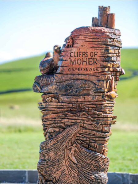 Im Bild ist eine Holzskulptur für die Cliffs of Moher Experience zu sehen. Dahinter sieht man die grüne Landschaft Irlands, die dazu einlädt durchwandert zu werden.