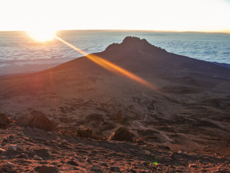 Die Sonne geht hinter dem Gipfel des Kilimandscharo auf. Sie taucht gerade aus dem Wolkenmeer, das hinter dem Gipfel zu sehen ist, auf.