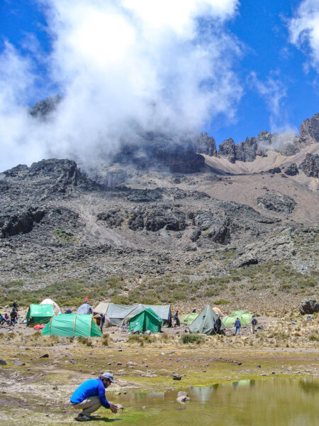 Im Vorderung hockt ein Wanderer an einem Bergsee in Tansania. Im Hintergrund sieht man ein Zeltcamp auf dem Weg zum Kilimanjaro.