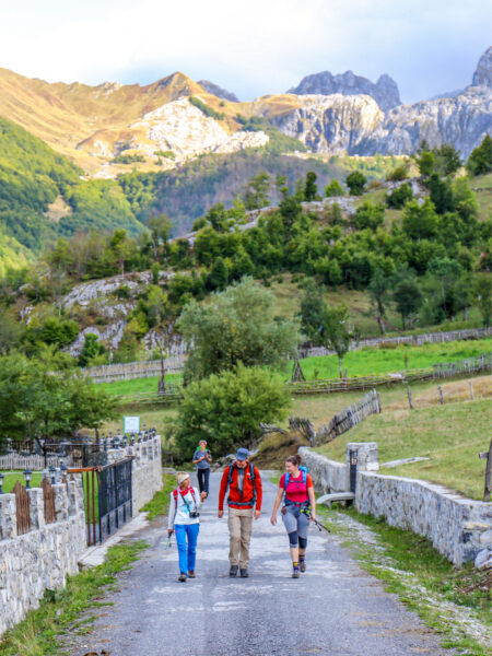 Drei Person auf Wanderreise durch den Balkan folgen einer Straße in Albanien oder Montenegro. Im Hintergrund ragen mehrere Gipfel des Berglands im Balkan in den Himmel auf.