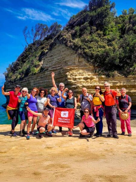 Eine Reisengruppe steht am Strand der griechischen Insel Korfu. Sie sind Teil einer Wanderreise des Reiseveranstalters Weltweitwandern, dessen Flagge sie auch zwischen sich halten.