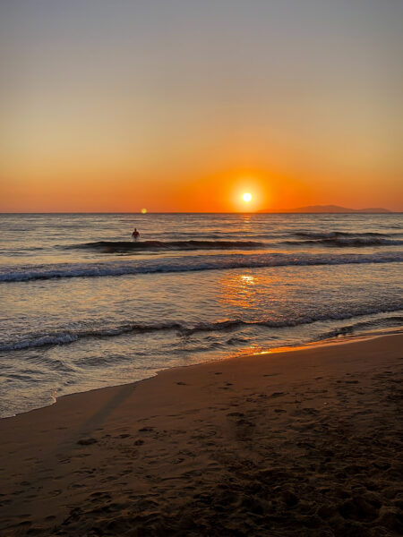 Die Sonne geht auf der Insel Korfu unter. Sie verschwendet quasi im ionischen Meer, während man im Vordergrund einen der schönen Strände der Region sieht.