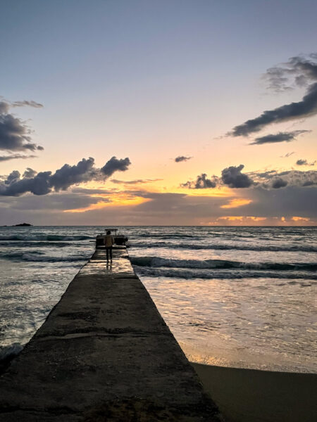 Eine Person steht auf einem Steg an der Küste von Korfu und schaut hinaus auf die Ionische See. Das Bild wurde beim Wanderurlaub der Person kurz nach Sonnenuntergang aufgenommen.