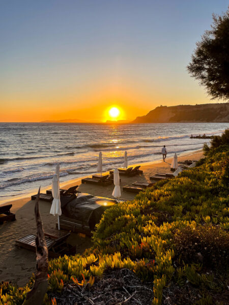 Die Sonne geht an der Westküste von Korfu unter. Im Vordergrund sieht man einen der Strände Insel sowie Schirme und Strandliegen.