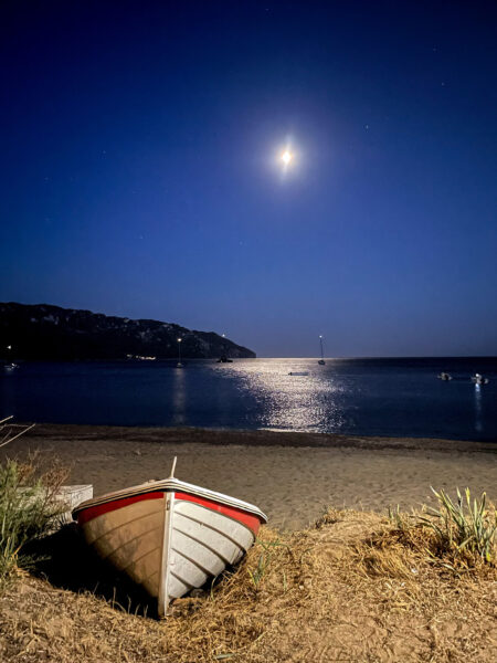 Ein Foto eines Ruderboots, das auf einem der Strände von Korfu liegt. Im Hintergrund sieht man den Nachthimmel und der Mond, der über der Szene hängt und für eine romantische Stimmung für eine Wanderreise auf der Insel sorgt.