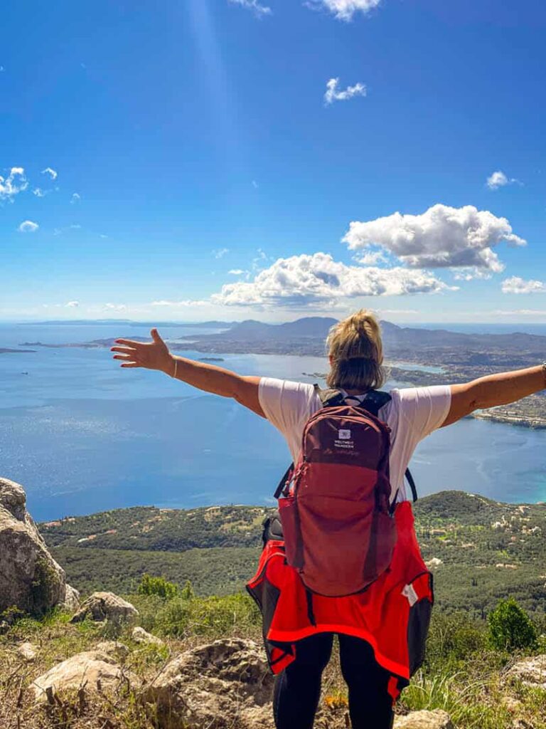 Unsere Local Guide aus Griechenland steht am Ausblick des Pantokratoras auf der griechischen Insel Korfu, auf der sie Gäste während einer Wanderreise herumführt. An ihr vorbei sieht man die ionische See.