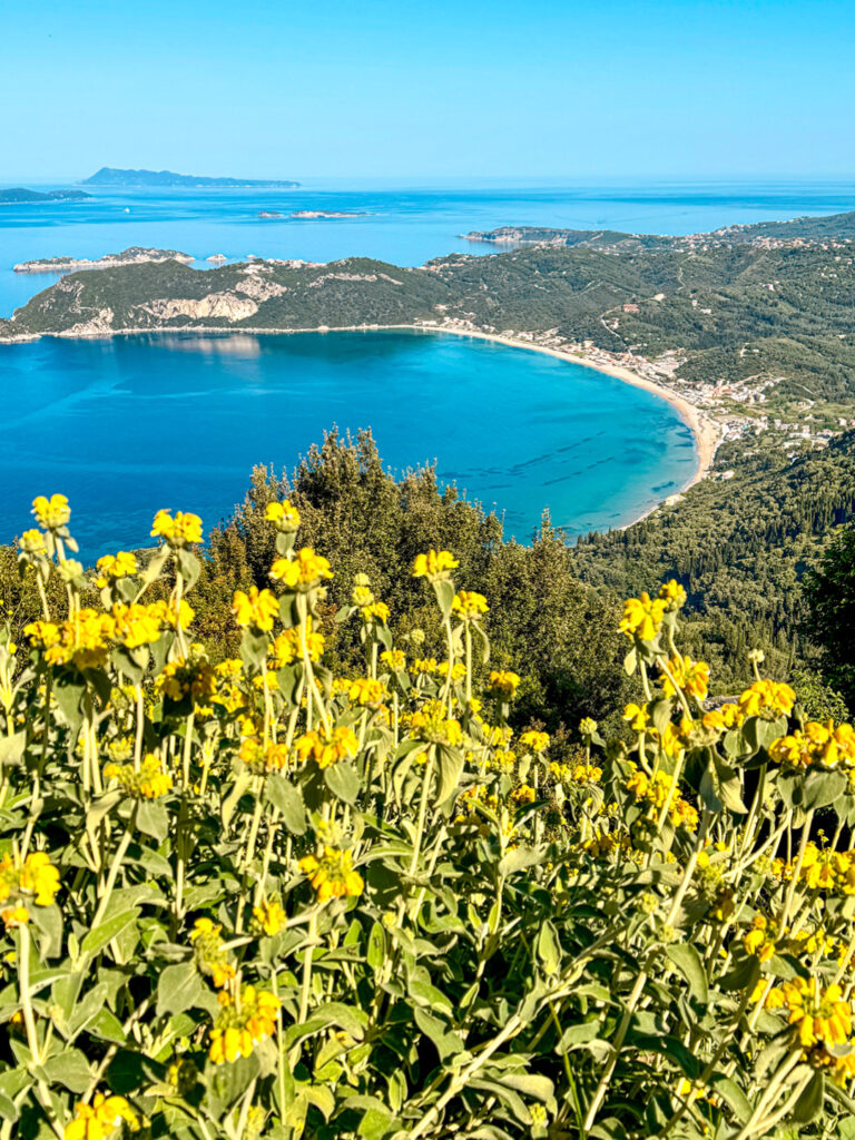 Das Bild zeigt die Bucht von Agios Georgios auf der Insel Korfu. Das klare Wasser des ionischen Meers strahlt dunkelblau und türkis zur Kamera herauf. Das Foto wurde von einem erhöhten Punkt auf einem Wanderweg aufgenommen.