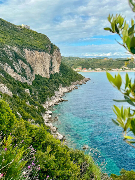 Man sieht die Buch von Agios Georgios zwischen den Blättern eines Olivenbaumes hindurch. Der Anblick offenbart die steilen Klippen der Bucht sowie die tiefen Wasser an der Küste Korfus im ionischen Meer.