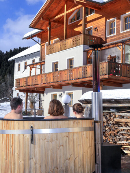 Drei Personen auf Wanderreise sitzen in einem kleinen, hölzernen Hot Tub vor einer Hütte im Lesachtal.