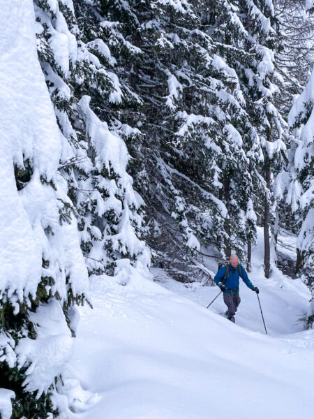 Ein Man kommt einen eingeschneiten Hang zwischen zwei Nadelbäumen hoch. Er trägt Schneeschuhen, die er für die Überquerung der winterlichen Landschaft braucht.