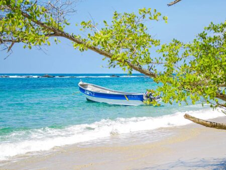 Ein kleines, blauweißes Boot liegt vertäut am Sandstrand im Tayrona-Nationalpark. Der Ast eines Baums ragt quer durch das Bild und bietet Schatten für die Reisenden von Weltweitwandern auf ihrer Wanderreise. Die Wellen brechen hier sanft an der kolumbianischen Küste.