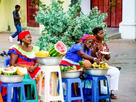 Drei kolumbianische Frauen in farbenfroher Kleidung sitzen auf Stühlen und verkaufen Obst, unter anderem an Wanderreisende der kleinen Gruppe von Weltweitwandern.