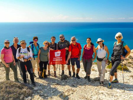 Eine kleine Reisegruppe steht gemeinsam am Höhepunkt einer ihrer Wanderungen auf der Mittelmeerinsel Zypern. Hinter ihnen sieht man das Mittelmeer und zwei Reisende halten zwischen sich die Flagge des Reiseveranstalters Weltweitwandern.