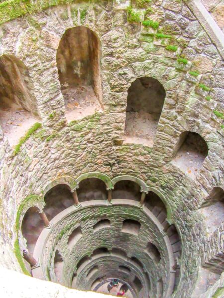 Eine tief gehende Wendeltreppe von oben betrachtet. Mehrere Fenster schauen von der weitläufigen Treppe auf einen offenen inneren Bereich. Moss bewächst die alten Steine einer alten Burganlage in Sintra in Portugal. Am tiefsten Punkt steht eine kleine Gruppe, die auf Wanderreise hierhergekommen ist.