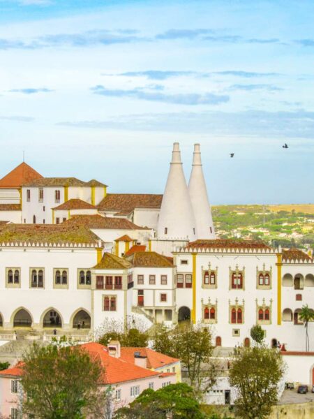 Man sieht einen Ausschnitt des Nationalpalasts in Sintra in Portugal. Die beiden konischen Küchenschornsteine des früheren maurischen Gebäudes ragen in den Himmel hinauf, die Dächer des Palasts und der umliegenden Häuser erstrahlen rot. Im Hintergrund kann man die Landschaft Portugals erkennen.
