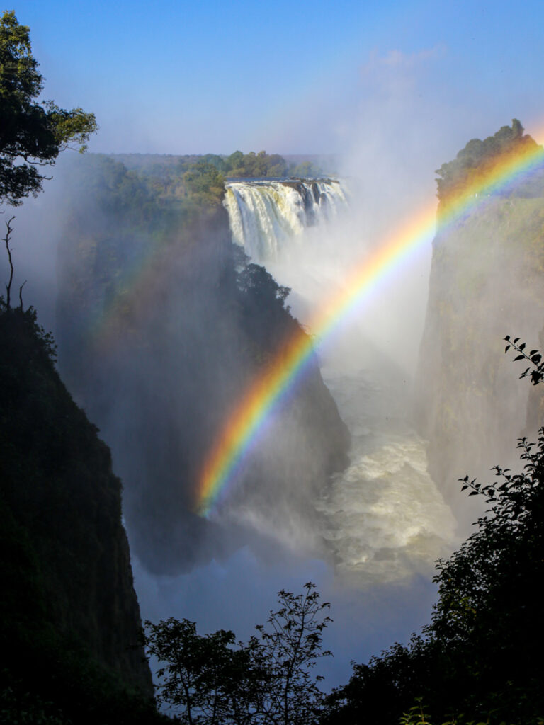 Ein Regebogen erstrahlt im Sprühnebel vor den Victoria Falls. Das UNESCO Weltnaturerbe der Wasserfällen in Simbabwe ist eines der vielen Naturhighlights bei einer Reise mit Weltweitwandern im Süden von Afrika.
