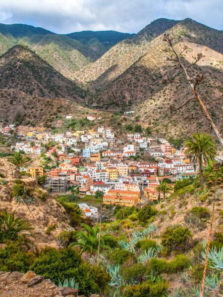 Am Bild ist die Ortschaft Vallehermoso auf der Insel La Gomera zu sehen. Zwischen der Person auf Wanderreise, die das Bild aufgenommen hat und der Ortschaft liegt eine karge Landschaft mit verzeinzelten Sträuchern, hinter der Ortschaft ist eine Gebirgslandschaft auszumachen.