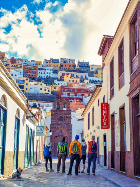 Vier Personen auf Wanderreise stehen in einer Gasse in San Sebastian auf der kanarischen Insel La Gomera. Sie stehen mit dem Rücken zur Kamera, an ihnen vorbei sieht man die bunten Häuser der größten Ortschaft auf der Insel, die sich am Hang hinaufarbeiten.