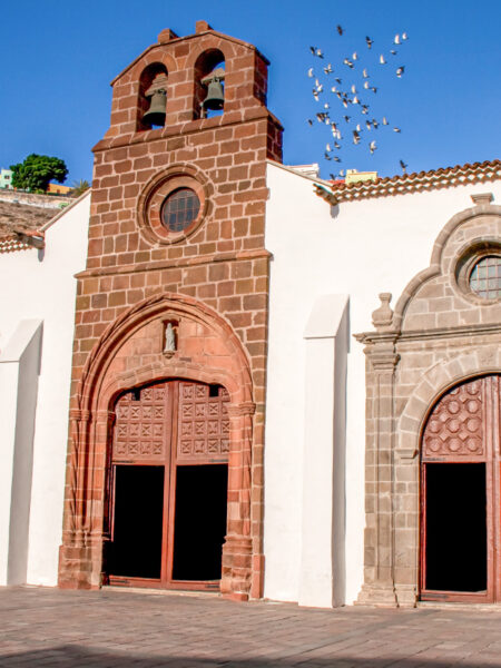 Im Vordergrud des Bildes ist die Iglesia de Nuestra Señora de la Asunción, die Kirche Mariä Himmelfahrt, in San Sebastián auf La Gomera zu sehen, dahinter fliegt ein Schwarm Vögel im Himmel.