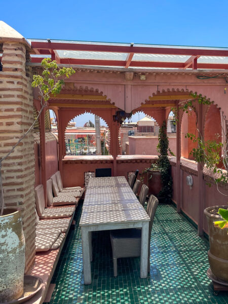 Auf einem Dach in Marrakesch stehen ein Tisch und mehrere Stühle, zu Teilen auch unter einem Überbau, der Schatten spenden soll.