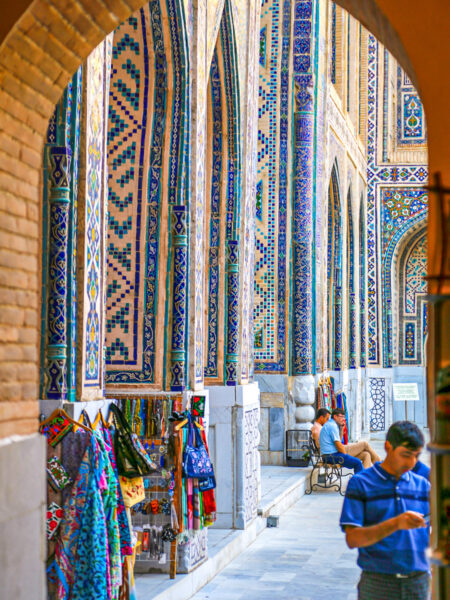 Man sieht einen Torbogen, durch den hindurch man die Shakhi-Zinda-Nekropole in Samarkand in Usbekistan erkennen kann. Im Vordergrund steht ein Einheimischer Mann.