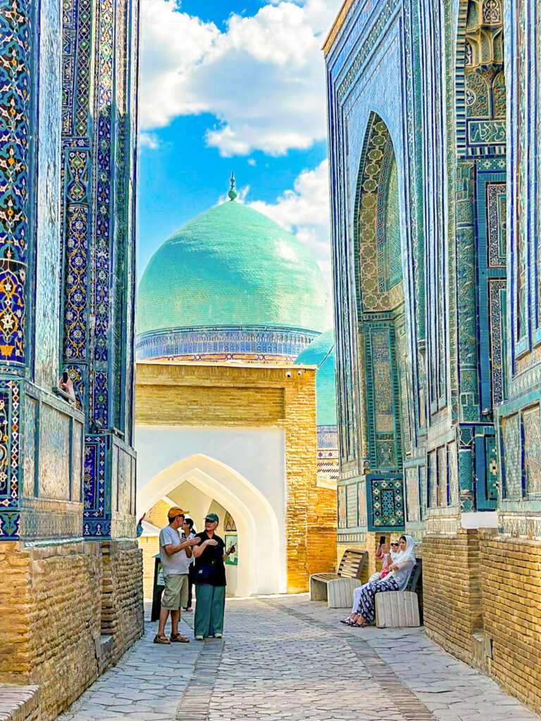 Man sieht eine Gasse in der Shah-i-Zinda-Nekropole in Samarkand. Mehrere Wanderreise stehen hier zwischen den farbenprächtigen Gebäuden und bekommen Infos zur Kultur und Tradition von Usbekistan.