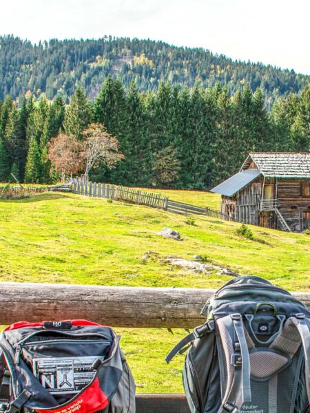 Zwei Wanderrucksäcke lehnen an einem Holzzauen. Dahinter sieht man angeschnitten eine Holzhütte und einen herbstlichen Nadelwald im Lesachtal in Kärnten.