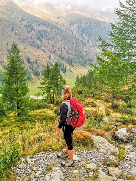 Eine Frau mit Wanderrucksack steht am Rande eines Weges im Lesachtal und schaut zwischen Nadelbäumen hindurch den Hang hinab. Der Rucksack ist rot wie das Logo von Wetweitwandern darauf.