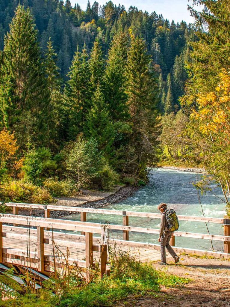 Ein Person wandert über eine Holzbrücke über einen Fluss, vermutlich die Lesach in Känrten. Das Bild ist eingerahmt von herbstlichen Wäldern.