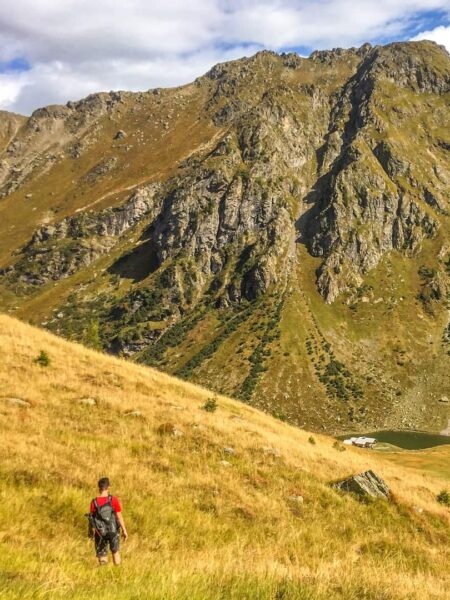 Man sieht eine Wiese im Herbst im Lesachtal. Im Hintergrund sieht man die kargen Wände eines Berges, klein im Vordergrund steht eine Person mit einem Wanderrucksack und einem roten T-Shirt von Weltweitwandern. Man sieht die Person von hinten.